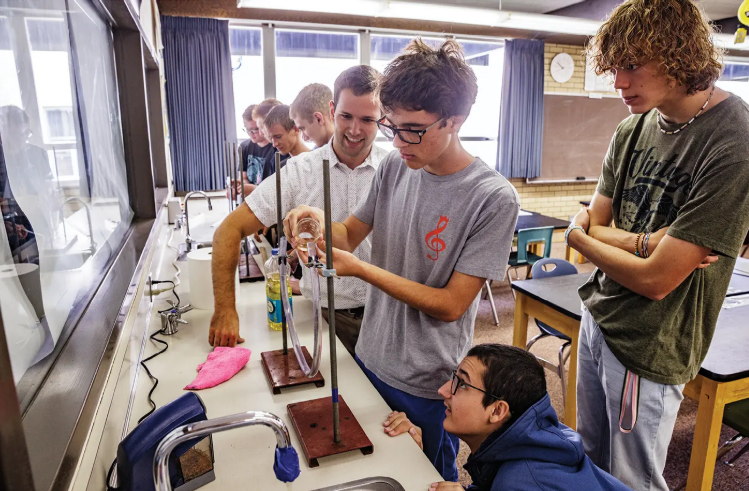 A science teacher guiding pupils through a chemistry demonstration with a clamp stand and gas delivery tube